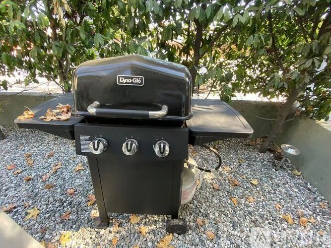 A Dyna-Glo grill is on a gravel surface surrounded by leaves.