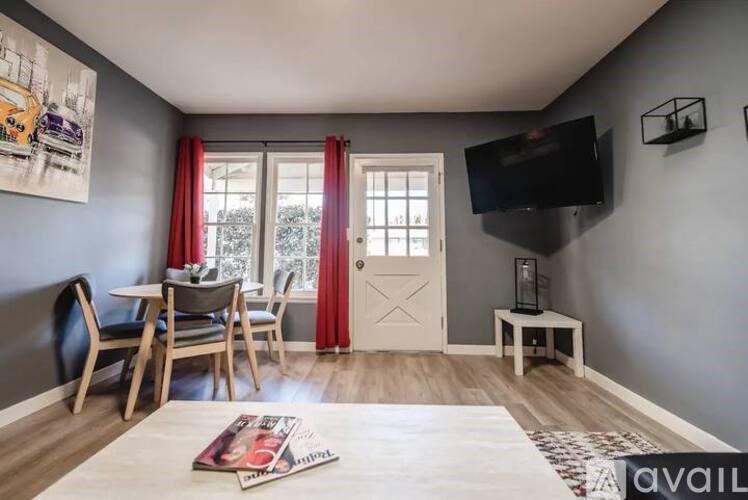 A living room with a white door, a grey sofa, a wooden table and chairs, a flat screen TV, and a painting on the wall.