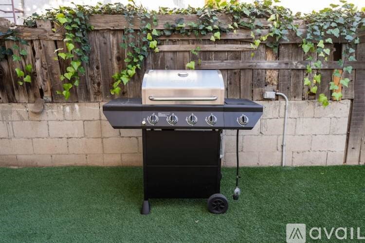A portable gas grill is on a green surface with a wooden fence and ivy in the background.