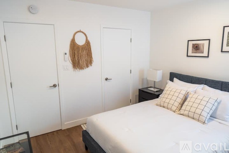 A bedroom with a bed, two pillows, a woven bag on the wall, and a lamp on a nightstand.