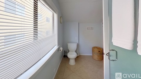 A bathroom with a toilet, a window with blinds, and a basket on the floor.