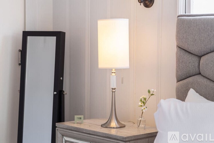 A bedroom with a grey headboard, a bedside table with a lamp, a vase with flowers, and a mirror.