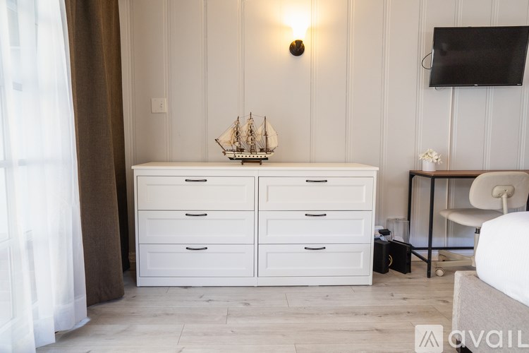 A white dresser with a ship model on top is in a room with a white wall and a window with curtains.