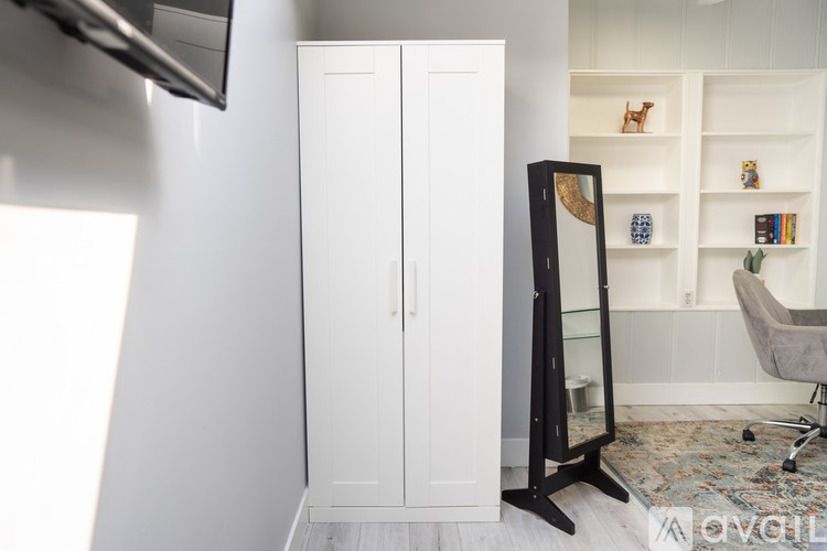 A tall white wardrobe stands in a room with a chair and a shelf with decorations.