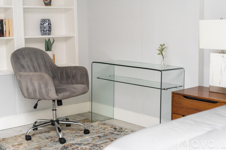 A grey chair is in front of a glass table and a shelf with books and vases.