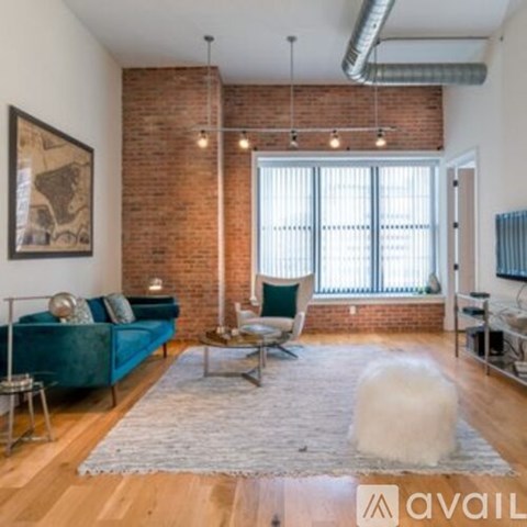A living room with a blue couch, a white fluffy ottoman, and a brick wall.