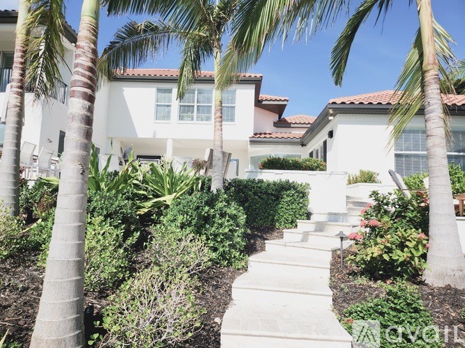 A white house with a red tile roof is surrounded by green plants and trees.