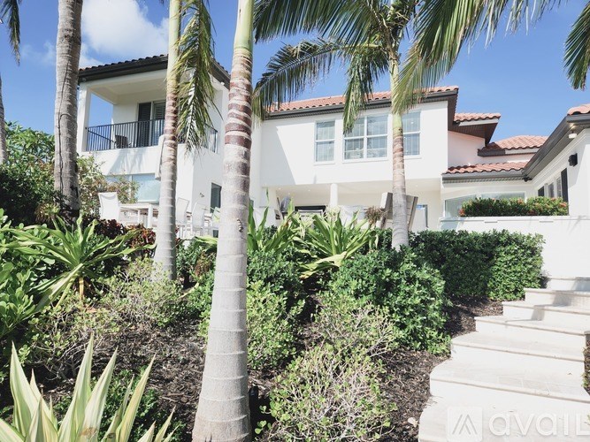 A white house with a red tile roof is surrounded by green plants and trees.