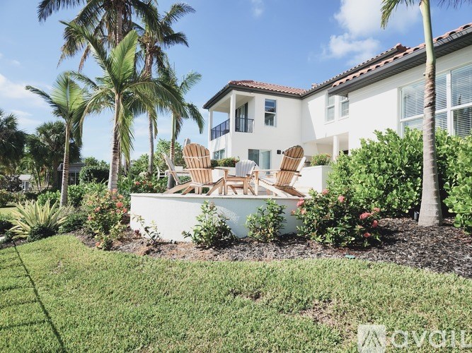 A white house with a red tile roof and a balcony is surrounded by palm trees and a garden with chairs.