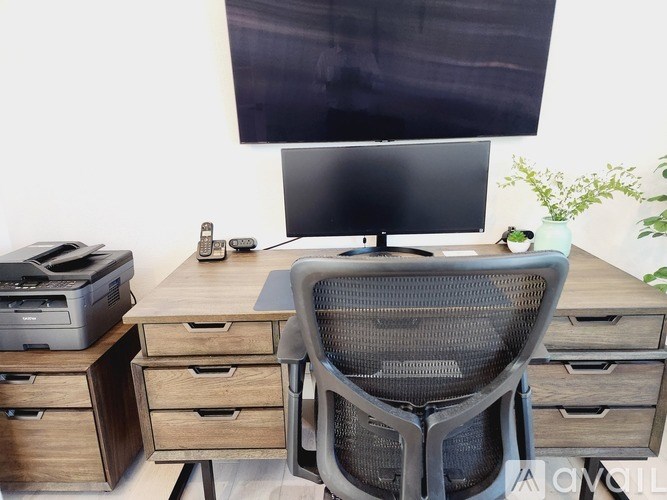 A black computer monitor is on a desk with a black chair and a printer.