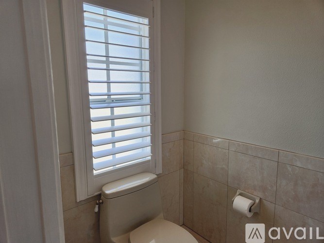 A bathroom with a toilet, a window with blinds, and a roll of toilet paper.