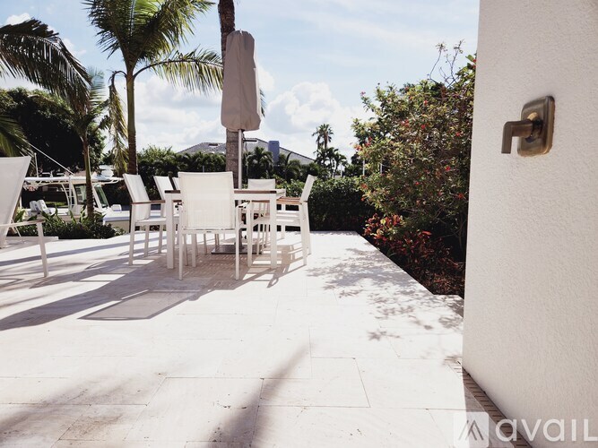 A white patio with chairs and a table is surrounded by greenery.