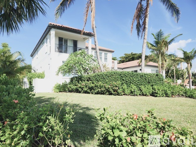 A white house with a red tile roof surrounded by greenery and palm trees.