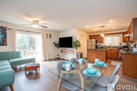 A living room with a glass table and chairs, a sofa, a television, and a kitchen area in the background.