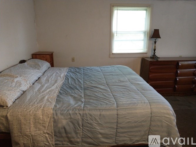 A bed with a blue and white comforter is in the foreground of an image with a window and lamp in the background.