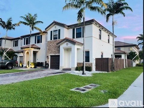 A house with a white exterior and a brown roof is surrounded by a green lawn and palm trees.