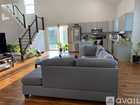 A grey couch is in the middle of a living room with a kitchen in the background.