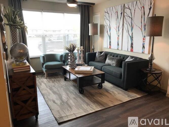 A living room with a black couch, a blue chair, a wooden coffee table, and a large window with blinds.