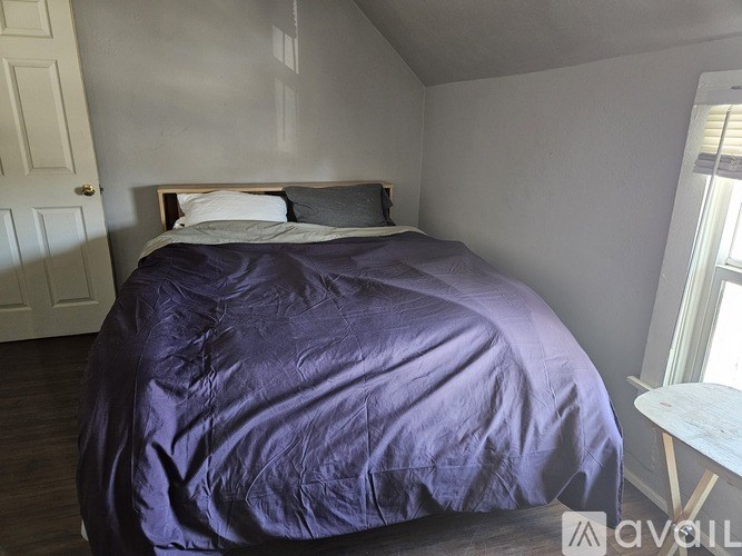 A bed with a purple comforter and two pillows sits in a room with a white door and a window.
