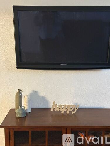 A flat screen TV mounted on a wall above a wooden table with a bottle and a sign that says "happy" on it.