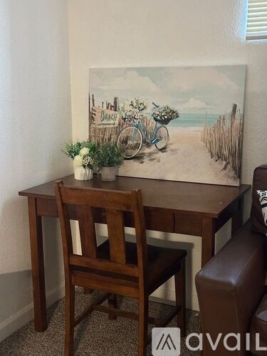A painting of a beach scene with a bicycle is hanging on the wall above a wooden desk with a chair.