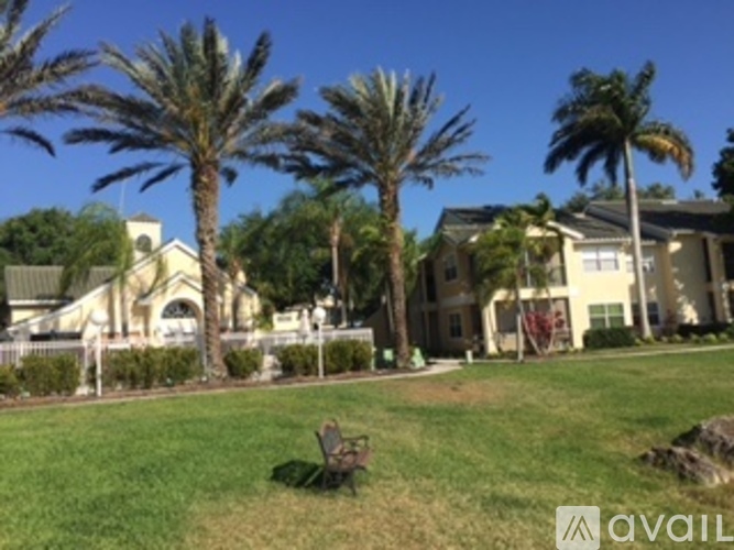 A bench sits in the foreground of a grassy area with palm trees and houses in the background.