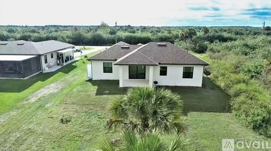 A white house with a brown roof is surrounded by greenery and has a sign that says "available".