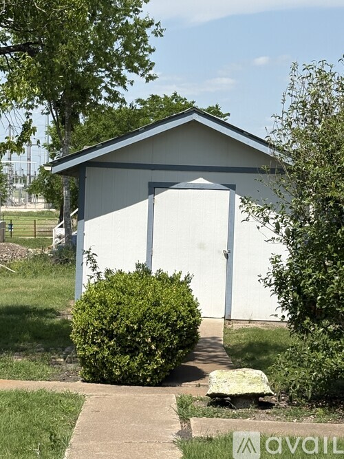 A small white building with a grey roof and a white door is surrounded by greenery.