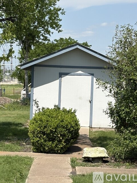 A small white building with a grey roof and a white door is surrounded by greenery.