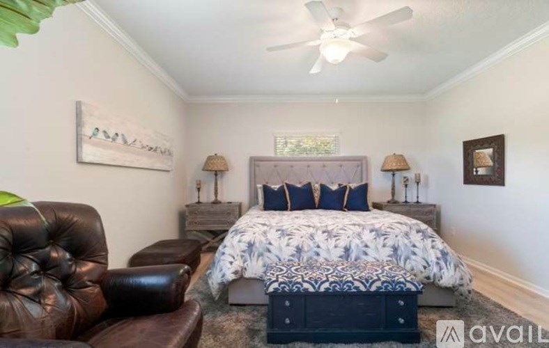 A bedroom with a leather chair, a bed with a blue and white comforter, and a ceiling fan.