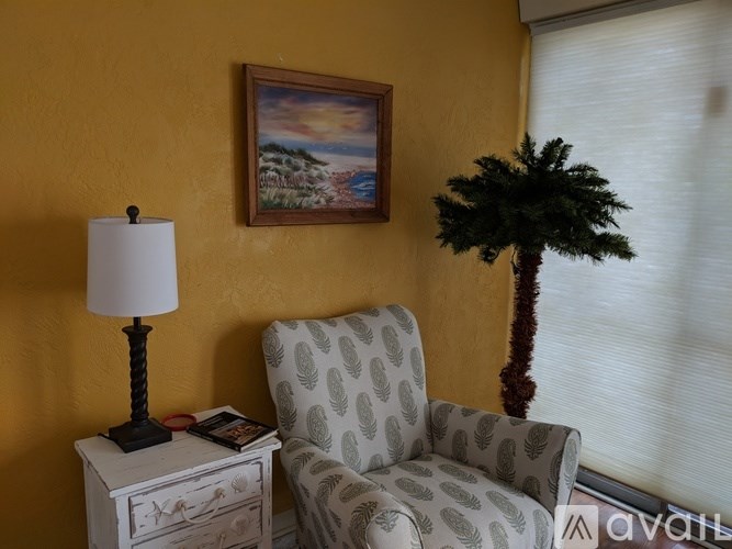 A chair with a patterned cushion sits next to a lamp and a painting on a yellow wall.
