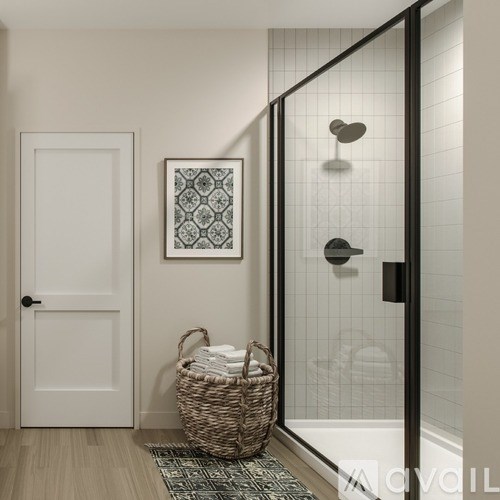 A bathroom with a white door, a framed picture on the wall, a basket with towels and a glass shower stall.