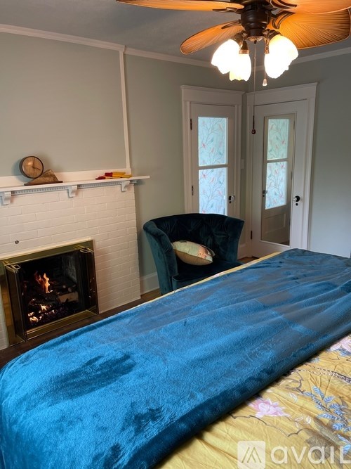 A bed with a blue blanket is in the foreground of a room with a fireplace and a ceiling fan.