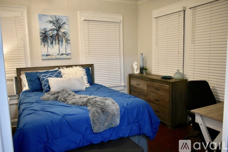 A bedroom with a blue bedspread and a painting of palm trees on the wall.