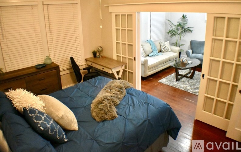 A bedroom with a bed covered in a blue comforter and two pillows, a desk with a chair, and a couch in the background.