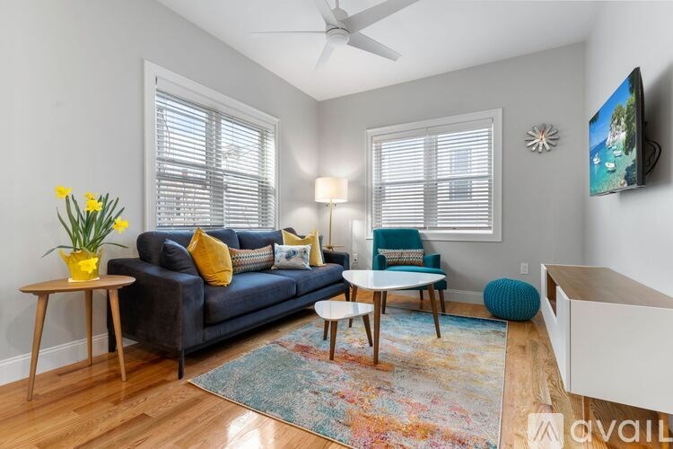 A living room with a blue couch, a yellow vase with flowers, a blue chair, a white side table, a wooden coffee table, a colorful rug, a ceiling fan, a flat-screen TV, and a white cabinet