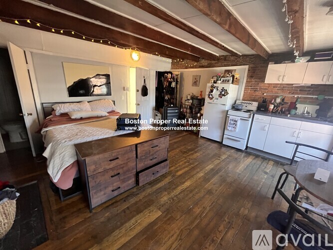 A bedroom with a bed, dresser, and a kitchen area in the background.