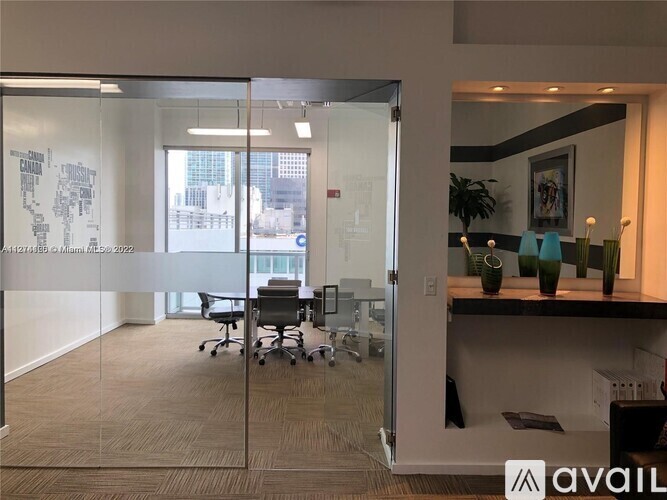 A glass wall separates two office spaces, one with a desk and chairs and the other with a shelf and vases.