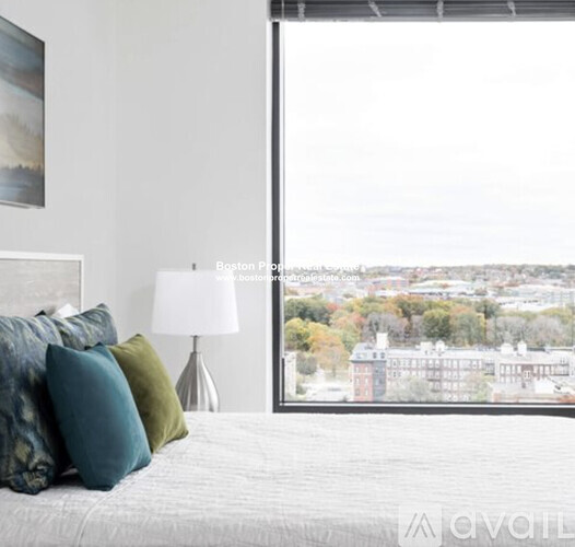 A bedroom with a bed, pillows, a lamp, and a window overlooking a cityscape.