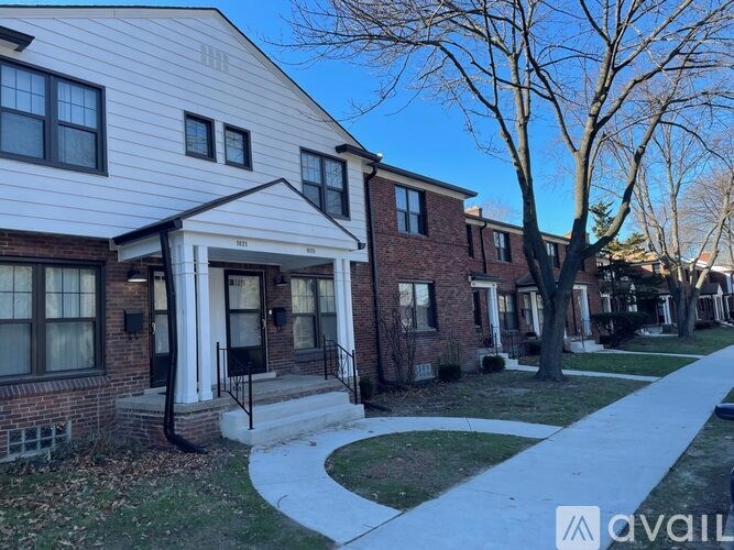 A white house with a black door and windows is in front of a tree and other houses.