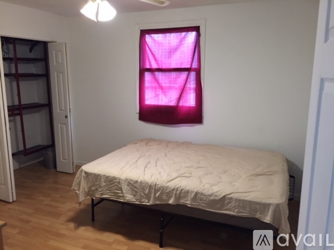 A bedroom with a bed, a window with a pink curtain, and a wooden floor.
