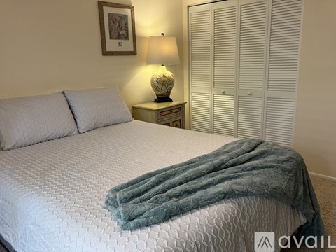 A bed with a white comforter and a grey blanket is in the foreground with a lamp and picture on the wall.