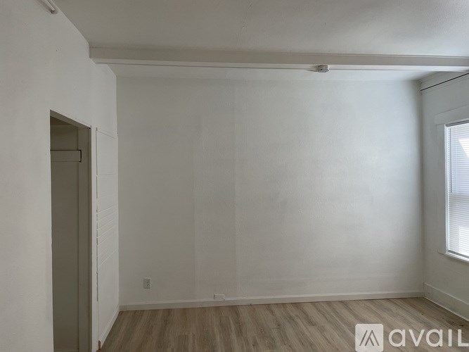 A room with wooden flooring and white walls, with a door on the left and a window on the right.