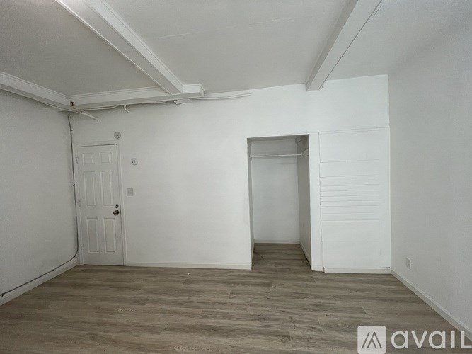 A room with wooden flooring and white walls, with a door on the left and a partially visible door on the right.