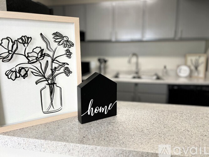 A framed drawing of flowers sits on a countertop next to a small black house-shaped sign that says "home.".