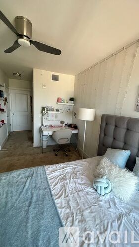 A bedroom with a bed, a desk with a chair, a ceiling fan, and a wall hanging.