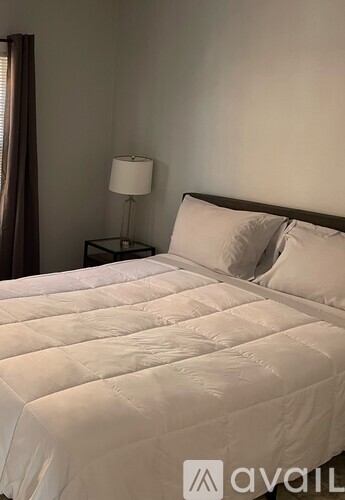 A bed with a white comforter and two pillows sits in a room with a lamp and curtains.