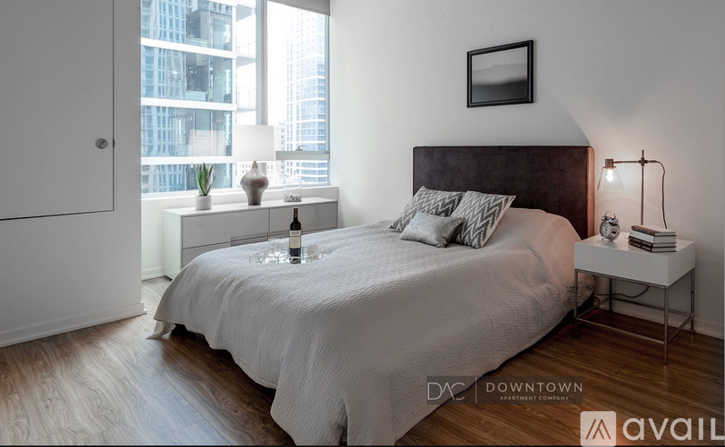 A bedroom with a large bed, a nightstand with a lamp, and a window overlooking a cityscape.