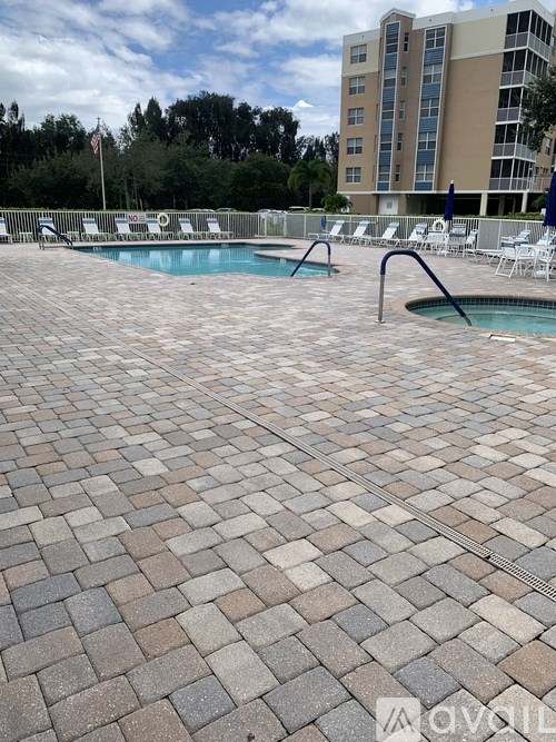 A pool surrounded by a brick patio with chairs and a building in the background.