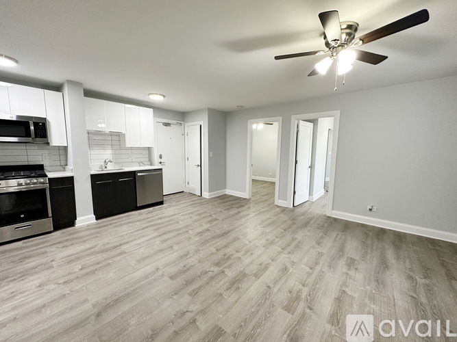 A spacious living room with a ceiling fan and light fixture, hardwood flooring, and a kitchen area with stainless steel appliances.